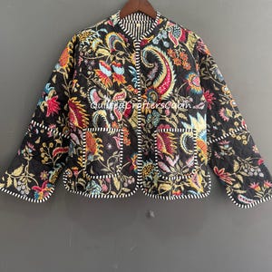 May include: A black quilted jacket featuring a vibrant multi-colored floral and paisley print. The garment has a round neckline, long sleeves, and two front pockets, all finished with a black and white striped trim. Text reads QuiltedCraftersCabin.