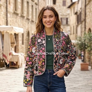 May include: A quilted jacket featuring a black base with a vibrant floral pattern in shades of pink, yellow, and white. The jacket has long sleeves, an open front, and a decorative stitched trim along the edges. It is worn over a dark green top and blue denim jeans.