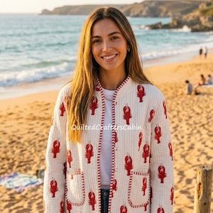 May include: A cream-colored quilted jacket featuring a repeating pattern of red lobsters. The jacket has a round neckline, long sleeves, two front patch pockets, and red and white striped trim along the edges and the front opening.