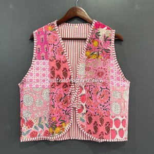 May include: A pink quilted vest with a patchwork design featuring floral and geometric patterns. The vest has a striped lining and is trimmed with white piping. The text "QuiltedCraftersCabin" is visible on the vest.