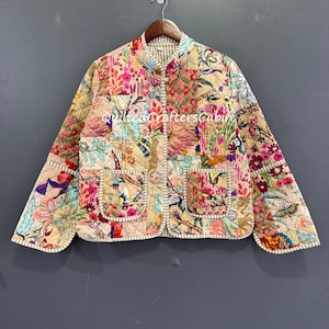 May include: A quilted jacket with a vibrant floral patchwork design. The jacket features a beige background with colorful patches of flowers, birds, and leaves. It has two front pockets and a striped lining. The text "Quilted Crafters Cabin" is visible.