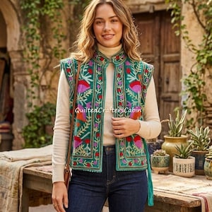 May include: A sleeveless waistcoat in deep green featuring vibrant floral embroidery in shades of pink, orange, and purple. The waistcoat has a high collar, a straight front opening, and is worn over a cream-coloured long-sleeve top and dark blue jeans.