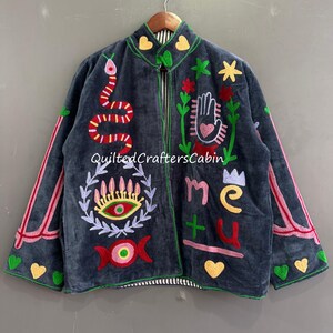 Op de afbeelding: Donkergrijze jas met kleurrijke borduursels, waaronder een slang, een hand, een oog en harten. De jas heeft een ritssluiting en de letters "me" en "u". De merknaam "QuiltedCraftersCabin" is ook zichtbaar.