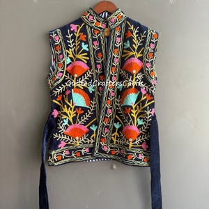 Puede incluir: Un chaleco sin mangas de color azul oscuro con bordados florales multicolores en tonos naranja, rosa y azul claro. La prenda tiene cuello alto, frente abierto y largas cintas de tela azul oscuro en la cintura. El texto dice QuiltedCraftersCabin.