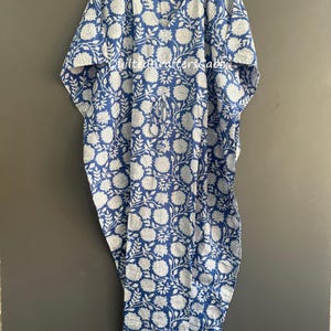 Op de afbeelding: Een blauw-witte kaftan met een herhalend bloemenpatroon van bladeren en bloesems. Het kledingstuk heeft een V-hals, wijde mouwen en een tailleband met trekkoord. De tekst QuiltedCraftersCabin is in het wit op de borst gedrukt.