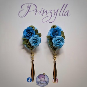 Könnte beinhalten: Ein Paar handgefertigte Ohrringe mit blauen Rosenblumendesigns. Jeder Ohrring hat eine hellblaue ovale Basis mit drei blauen Rosen und grünen Blättern. Goldfarbene, längliche Tropfenanhänger hängen darunter. Der Markenname "Prinzylia" ist sichtbar.