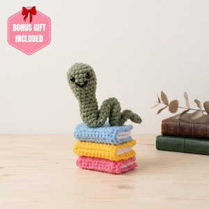 Può includere: Un verme all'uncinetto verde con occhi a perla neri e un sorriso ricamato, appoggiato su una pila di tre piccoli libri rettangolari all'uncinetto in azzurro, giallo e rosa. Un badge esagonale rosa nell'angolo riporta BONUS GIFT INCLUDED.