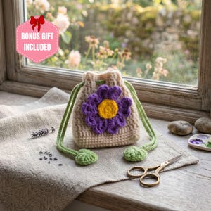 May include: A beige crochet drawstring pouch featuring a large purple flower with a yellow center on the front. The pouch has green drawstrings with small green leaf-shaped ends. A pink hexagonal sign reads BONUS GIFT INCLUDED.