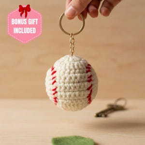 May include: A cream-coloured crocheted baseball keyring featuring red yarn stitching details. The baseball is attached to a gold-toned metal split ring by a short chain. A pink hexagonal graphic in the upper left corner displays the text BONUS GIFT INCLUDED.