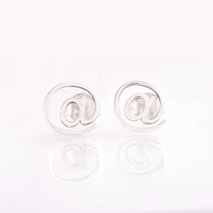 Op de afbeelding: Een paar zilverkleurige oorstekers, elk in de vorm van het "@"-symbool. De oorbellen staan tegen een witte achtergrond, met een subtiele reflectie eronder.