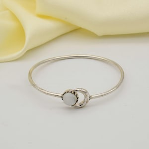Könnte beinhalten: Ein silberfarbenes Armreif-Armband mit einem Mond-Design und einem runden, weißen Edelstein in der Mitte. Das Armband ist schlicht und elegant. Der Hintergrund ist ein weicher, cremefarbener Farbton. Der Durchmesser beträgt ca. 6 cm.
