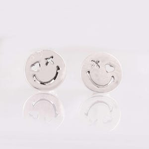 May include: A pair of silver-colored stud earrings. Each earring features a smiley face design with a heart-shaped eye and a cross-shaped eye. The earrings are round and have a reflective surface.