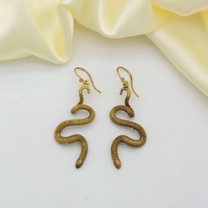 May include: A pair of gold-colored snake earrings. The earrings are shaped like snakes with textured, scale-like detailing. The snake earrings have a hook closure and are displayed on a white surface with a yellow fabric in the background.