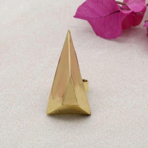 May include: A gold-colored ring with a geometric pyramid design. The ring features a solid, triangular shape with a pointed top and a band for the finger. The ring is made of a shiny, metallic material.
