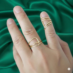 Puede incluir: Dos anillos dorados se muestran en dedos sobre un fondo verde. Un anillo presenta un diseño geométrico con forma triangular, mientras que el otro tiene un diseño abierto y en capas. Los anillos son de metal brillante.