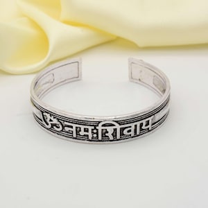 May include: Silver-toned cuff bracelet with engraved Sanskrit text. The open-ended bracelet features a detailed design with a central band of text and decorative borders. The bracelet is displayed on a light background with a soft, yellow fabric.
