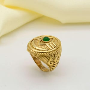 May include: A gold-colored ring with an eye-shaped design and a small green stone in the center. The ring has intricate detailing, including a rope-like border and decorative patterns on the sides. The ring is set against a soft, off-white background.