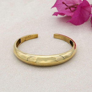 May include: A polished gold-colored cuff bracelet. The open-ended bracelet has a smooth, rounded design. The word "Iddo" is engraved on the inside of the bracelet. A pink flower is in the background.