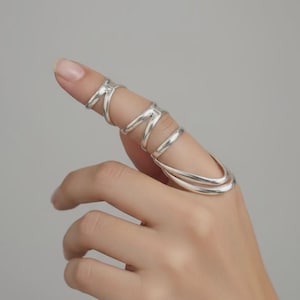 May include: A silver-colored finger ring with a unique design. The ring wraps around the finger with multiple bands and a curved, open design. The jewelry is displayed on a hand against a neutral background.