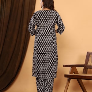 K&ouml;nnte beinhalten: Schwarzes und wei&szlig;es gemustertes Kurta- und Hosen-Set. Das Outfit hat ein sich wiederholendes Blumenmuster. Die Dreiviertel-&Auml;rmel-Kurta und die passende Hose werden mit goldfarbenen Abs&auml;tzen getragen. Der Hintergrund umfasst einen braunen Vorhang und einen Holzstuhl.