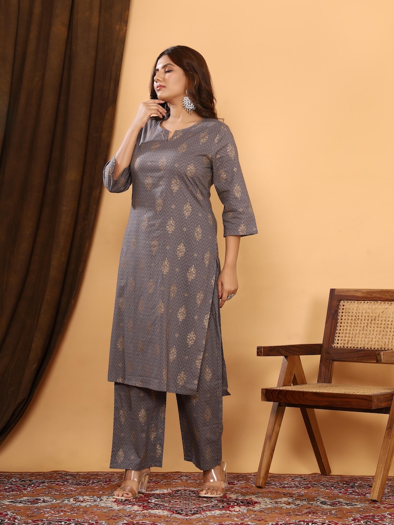 K&ouml;nnte beinhalten: Ein graues Kurta- und Palazzo-Set mit goldenem geometrischem Muster. Das Outfit hat einen V-Ausschnitt und Dreiviertel&auml;rmel. Die Palazzo-Hose hat ein weites Bein. Das Outfit wird mit transparenten Sandalen getragen.