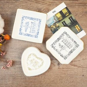 Puede incluir: Tres platos decorativos de cerámica con bordes dorados. Un plato cuadrado presenta un diseño floral azul y el nombre "Millie". Otro plato en forma de diamante tiene un diseño floral negro y el nombre "Henry". También se incluye un plato en forma de corazón.