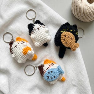 May include: Four handmade crochet cat keychains in various colors. One is black with a fish, one is white and black, and two are white, orange, and brown. Each keychain has a silver ring attached for keys.