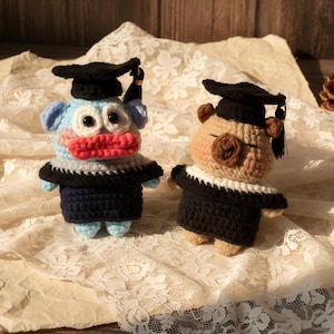 May include: Two handmade crochet figures, one blue and one tan, dressed in graduation attire. The blue figure has large eyes, a red mouth, and a black graduation cap. The tan figure wears glasses and a black cap. Both figures have black gowns.