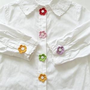 May include: A white long-sleeved shirt with a lace-trimmed collar and cuffs, featuring colourful crocheted flower appliques attached to the centre button placket and the cuffs. The flowers are red, pink, light purple, green, and yellow.