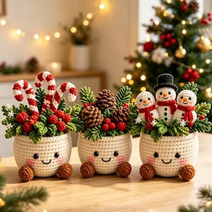 Könnte beinhalten: Drei gehäkelte cremefarbene Töpfe mit lächelnden Gesichtern, rosa Wangen und braunen Füßen. Der erste enthält rot-weiße Zuckerstangen und Beeren. Der zweite zeigt Tannenzapfen und Grün. Der dritte enthält drei kleine gehäkelte Schneemänner.