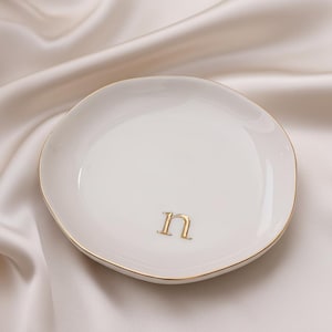 Puede incluir: Un plato pequeño y redondo de cerámica color blanco hueso con un borde irregular y orgánico. El centro del plato presenta una letra n minúscula en un acabado dorado metálico. El borde del plato también está acentuado con una línea dorada.