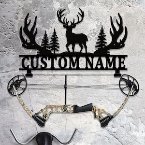 May include: Black metal wall art featuring a deer silhouette, trees, and the text "CUSTOM NAME". Below, a camouflage compound bow is mounted. The artwork is designed for hunters and outdoor enthusiasts.