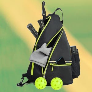 May include: Black and neon green pickleball bag with a single strap. The bag has a zippered main compartment and a front pocket. Inside the bag are two pickleball paddles, a gray towel, and a phone. Two neon green pickleballs are in front.