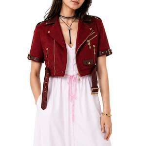 May include: A short-sleeved, cropped burgundy jacket with gold-tone zippers, a wide collar, and buckled straps on the sleeves and waist, worn over a light pink, button-down dress featuring a drawstring waist and a white lace-trimmed hem.