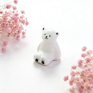 Könnte beinhalten: Kleine, weiße Keramik-Bärenfigur mit schwarzen Augen und Pfotenabdrücken. Der Bär sitzt mit ausgestreckten Beinen. Rosa Blüten sind auf beiden Seiten des Bären vor weißem Hintergrund angeordnet.