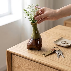 May include: A small, handcrafted ceramic vase with a brown and green glaze, holding a small bouquet of white and pink flowers. The vase is on a light wood surface, next to a small dish with rings and a key chain.