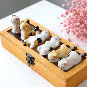 May include: A wooden box with a metal clasp holds six ceramic cat figurines. The cats are in different poses and are white, orange, and black. The box is light brown. The cats have pink accents.