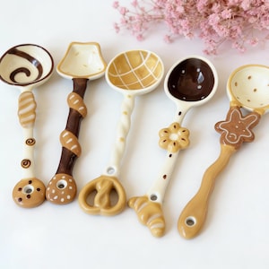 May include: A collection of five ceramic spoons with unique designs. Each spoon features a different handle and bowl shape, with brown, tan, and white colors. The handles are decorated with swirls, pretzels, and flower shapes. The spoons are arranged on a white surface.
