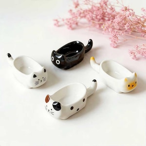 Op de afbeelding: Vier keramische katvormige schaaltjes in wit en zwart. De schaaltjes hebben een kleine komvorm met een kattengezicht aan de voorkant en een staart aan de achterkant. Eén schaaltje is effen zwart, de andere zijn wit met verschillende kattenmarkeringen.