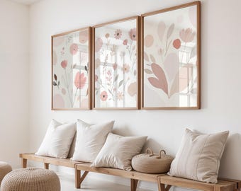 Op de afbeelding: Drie ingelijste bloemenkunstprints met licht houten frames. De prints tonen zachte roze en perzik bloemen en bladeren op een witte achtergrond. Daaronder staat een houten bank met witte kussens en een gevlochten mand.