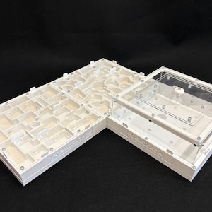 May include: A white plastic ant farm with a complex network of tunnels and chambers on the left side and a clear acrylic lid covering a separate section on the right. The word FORMICADA is embossed on the side of the white plastic structure.