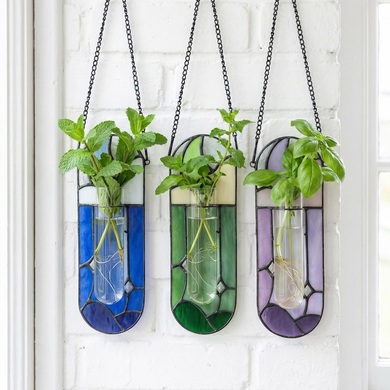 Puede incluir: Tres jardineras colgantes de vidrieras con cadenas de metal, cada una con un tubo de vidrio con una planta. Las jardineras presentan esquemas de color azul, verde y morado. Las plantas tienen hojas verdes y ra&iacute;ces visibles.