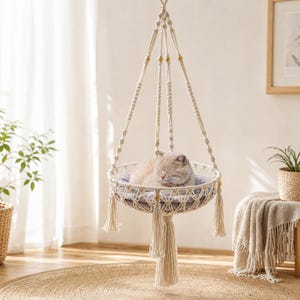May include: A cream-colored macrame hanging cat bed featuring intricate knotted rope patterns, wooden bead accents, and long decorative tassels. A light brown cat is curled up inside the circular basket, which is suspended by four braided ropes.