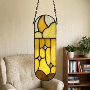 May include: A stained glass window hanging from a black chain. The window features yellow, white, and brown glass panels with a geometric design. The window is hanging in front of a beige wall.