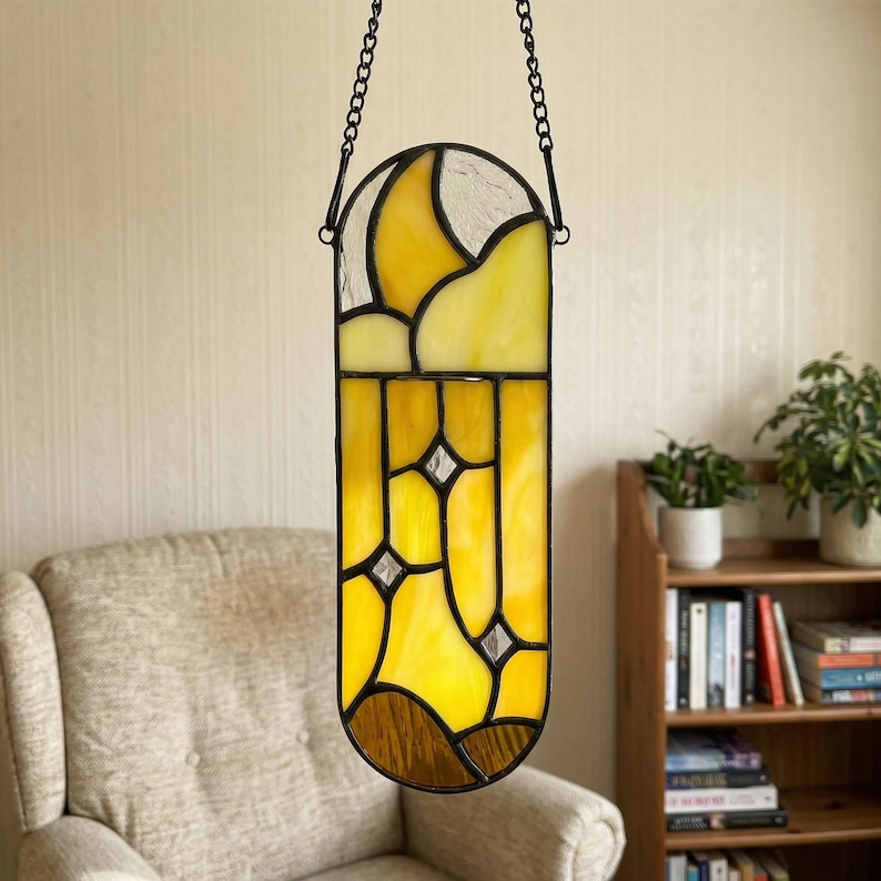 May include: A stained glass window hanging from a black chain. The window features yellow, white, and brown glass panels with a geometric design. The window is hanging in front of a beige wall.