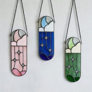 Puede incluir: Tres paneles de vidrieras en rosa, azul y verde, cada uno con un marco de metal negro y una cadena para colgar. Los paneles presentan un dise&ntilde;o celestial con estrellas y una luna creciente.