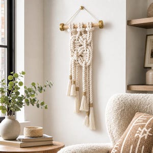 May include: A wall hanging made of thick off-white cotton rope knotted into a central geometric pattern. The rope is suspended from a light-colored wooden dowel with gold-toned metal end caps and hangs by a thin white cord from a gold hook.
