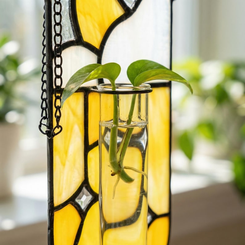 May include: A stained glass propagation station with a yellow and white design. A clear glass tube holds a plant cutting with green leaves. A black chain is attached to the stained glass panel.