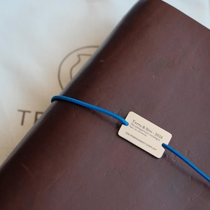 Puede incluir: Un diario de cuero marrón con una banda elástica azul y una pequeña etiqueta. La etiqueta dice "Terra & Slim - 2026" y "ON PERMANENT DISPLAY". Una bolsa de tela blanca está en el fondo.