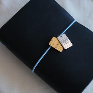 May include: A dark blue leather journal with a light blue elastic band closure. Two small rectangular tags hang from the band. One tag is gold-colored, the other is silver-colored. The journal is on a white surface.